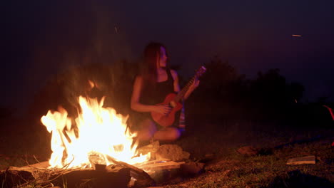 Mujer-Asiática-Tocando-La-Guitarra-Ukelele-Frente-Al-Fuego-En-La-Noche-Acampando-Al-Aire-Libre