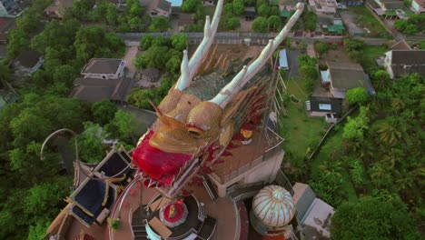 Aerial-shot-of-a-vibrant-dragon-sculpture-atop-a-temple-surrounded-by-lush-greenery,-daylight