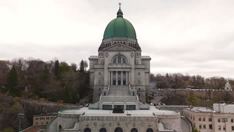 Aerial-front-view-of-Saint-Joseph's-Oratory-of-Mount-Royal,-Montreal,-drone-approaching-old-roman-Catholic-basilica-green-dome-revealing-stunning-cityscape-at-distance-during