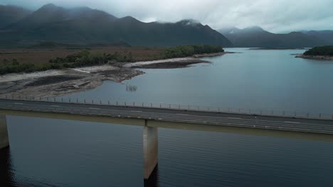 Vehicles-passing-on-a-bridge-over-lake-Burbury-with-beautiful-waterscape-and-landscape-of-mountains-in-Tasmania,-Australia