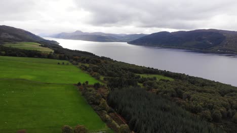 Aerial-View-Over-Green-Fields-River-Bank-Trees-With-Loch-Ness-In-The-Scottish-Highlands