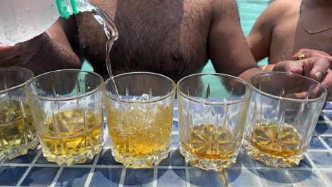 Closeup-view-of-pouring-of-liquor-in-glasses-beside-pool-during-daytime