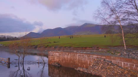 Excursionistas-Caminando-Cerca-De-Un-Lago-Tranquilo-En-El-Distrito-De-Los-Lagos-Al-Atardecer,-Como-Telón-De-Fondo-De-Montaña