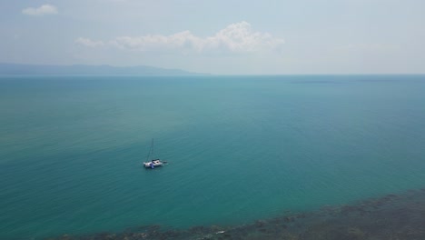 Sail-boat-floating-in-open-ocean-next-to-coral-reef---drone-view