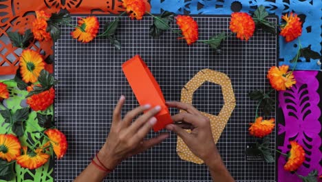 Hands-gluing-colorful-Day-of-the-Dead-box-on-vibrant-floral-backdrop,-top-view