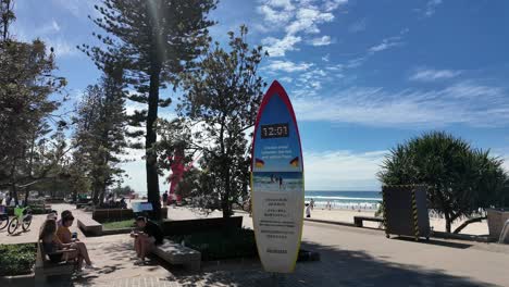 Gente-Relajándose-En-Una-Explanada-Cerca-De-Una-Playa-Alrededor-De-Un-Reloj-De-Tabla-De-Surf