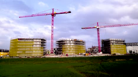 Sideways-aerial-looking-up-at-red-cranes-rising-above-three-residential-towers-under-construction-wrapped-in-yellow-scaffolding