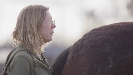 Tears-run-down-woman's-cheek-standing-behind-horse-during-equine-therapy,-profile