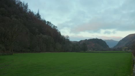 Dusk-settles-over-the-lush-green-fields-of-Lake-District,-with-hills-and-leafless-trees