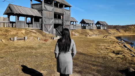 Back-View-of-Woman-Walking-Under-Wooden-Walls-of-Medieval-Fort-and-Town-on-Sunny-Day,-Slow-Motion