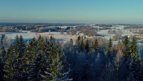 Aerial-winter-sunrise-over-snow-covered-forest-and-countryside,-cozy-rural-scenery