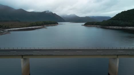Aerial-side-view-of-range-of-mountains-under-a-cloudy-day-in-Tasmania,-Australia-with-an-vacant-bridge-in-foreground
