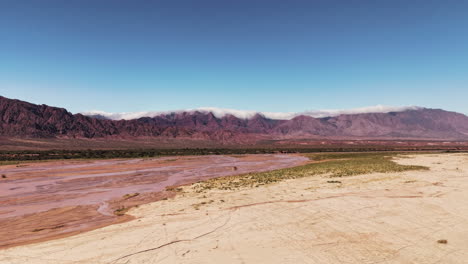 Hyperlapse-drone-footage-of-a-shallow-river-in-the-desert-with-the-Andes-mountain-range-in-the-background,-embraced-by-clouds
