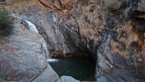 Water-cascading-into-a-serene-rock-pool-surrounded-by-rugged-cliffs-at-dusk,-time-lapse