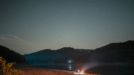Night-sky-over-serene-lake-with-stars,-small-campfire-on-shore,-mountains-in-background,-tranquil-atmosphere,-timelapse