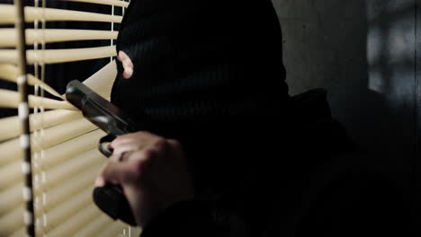 Close-up-of-criminal-peering-out-of-window-with-ski-mask-on-and-gun-at-night