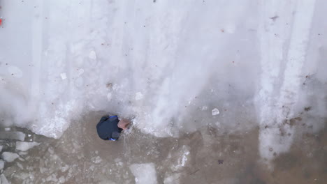 Ice-diver-in-drysuit-shatters-edge-of-lake-ice-to-open-entry-point,-top-drone