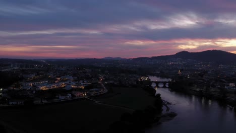 twilight-over-Barcelos-captures-town's-ambiance-as-night-approaches,-with-the-medieval-bridge-over-Cávado-River,-Portugal---aerial