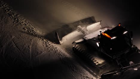 Snow-groomer-working-at-night-in-the-Dolomites,-Italy,-illuminating-snowy-slopes,-aerial-view