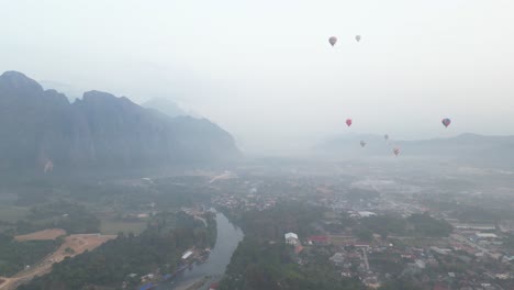 Toma-De-Drones-Del-Valle-Brumoso-Con-Globos-Aerostáticos-A-Lo-Lejos-En-Vang-Vieng,-La-Capital-De-Aventuras-De-Laos.