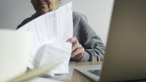 Foto-De-Primer-Plano-De-Una-Mujer-Mayor-Pasando-Por-El-Concepto-De-Pago-De-Facturas-En-Papel-Dentro-Del-Escritorio-Junto-A-La-Computadora-Portátil-De-Casa,-Concepto-De-Pago-Fuera-De-Línea-Versus-En-Línea