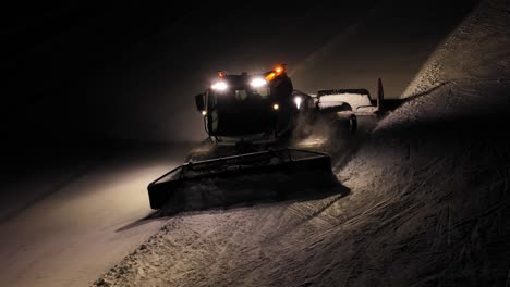 Snow-groomer-working-at-night-in-the-Dolomites,-Italian-Alps,-with-bright-lights-and-a-clear-trail-behind