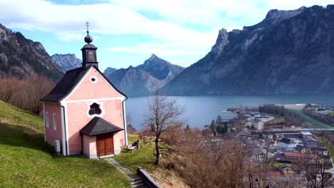 Drone-shot-pans-down-on-a-pink-chapel-on-Kalvarienberg-in-Ebensee,-with-Traunsee-lake-in-the-background,-peaceful-and-serene,-Upper-Austria