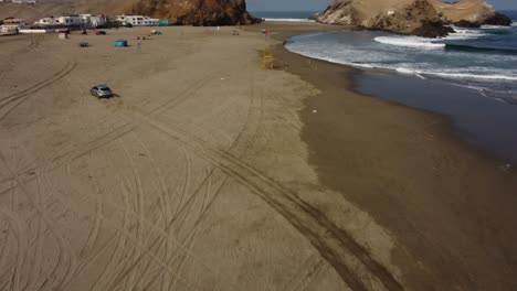 Toma-Aérea-De-Un-Automóvil-Alejándose-De-La-Orilla-De-Una-Playa-Dejando-Marcas-En-La-Arena