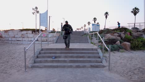 Un-Hombre-Con-Uniforme-Militar-Y-Portando-Un-Rifle-M4-Baja-Corriendo-Las-Escaleras-Hacia-La-Playa.