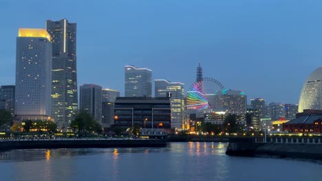 Evening-view-of-a-vibrant-city-skyline-with-illuminated-buildings-and-a-colorful-Ferris-wheel-reflected-in-the-water