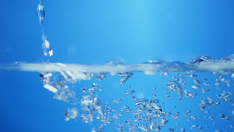 Pouring-into-a-half-filled-container-creating-waves,-ripples-and-droplets-of-water-against-a-blue-background