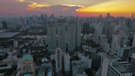 From-an-aerial-perspective,-Bangkok's-skyline-emerges,-showcasing-urban-living-against-a-sky-where-the-sun-peeks-through-crimson-hued-clouds,-creating-a-striking-scene