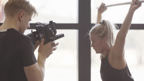 Guy-takes-video-of-blonde-female-fitness-influencer-squatting-barbell-above-head