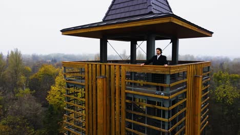 Man-in-black-coat-standing-in-watch-tower-above-the-forest,-aerial-establisher