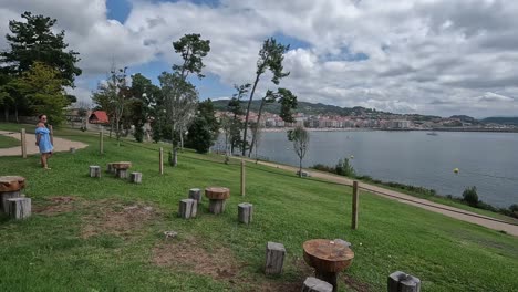 Woman-goes-out-to-enjoy-the-natural-grass-terrace-with-rustic-wooden-seats-and-tables-facing-the-sea-and-the-city-is-seen-in-the-background,-cloudy-day,-descriptive-shot