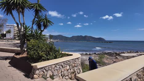 Cala-Bona,-Mallorca,-Spain-coastal-promenade-with-tourists,-palm-trees-and-hotels-overviewing-the-Mediterranean-Sea