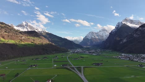 An-aerial-of-the-scenic-valley-of-Glarus-Nord,-Switzerland,-unveiling-a-charming-tableau-of-a-quaint-settlement-nestled-amid-snow-capped-peaks,-alpine-lifestyle-immersed-in-the-splendor-of-nature