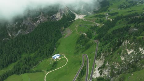 Aerial-perspective-of-Val-Gardena-pass,-drone-ascends-through-clouds,-revealing-winding-road-with-cars-below