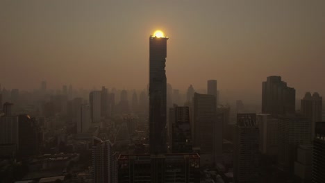 Aerial-view-of-Bangkok's-skyline,-the-atmospheric-scenery-reveals-urban-dwellings-amidst-a-sky-with-the-rising-sun-piercing-through-smoky-and-foggy-air