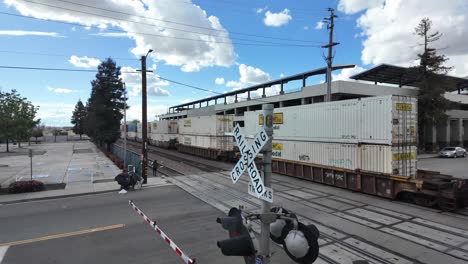 Freight-train-passing-a-crossing-in-Bakersfield,-sunny-day-with-dynamic-clouds,-barrier-down