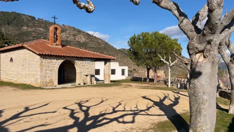 We-see-with-a-turn-of-the-camera-the-hermitage-of-San-Marcos-in-the-Barraco-Avila-is-made-of-stone-and-on-the-roof-is-the-small-bell-tower-it-has-a-dirt-square-with-trees-pruned-in-winter-Spain