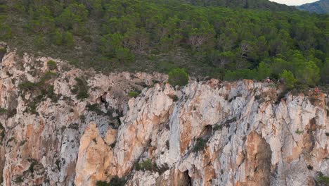 Hikers-rest-on-a-rocky-cliff-among-trees-with-a-view-of-the-green-landscape-during-sunset-in-ibiza