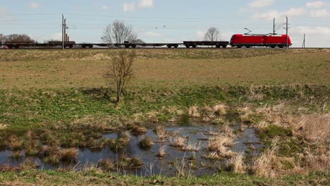 Red-electric-locomotive-pulling-freight-train-across-grassy-countryside,-sunny-day