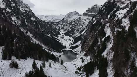 Tilt-Down-Reveal-of-Small-Lake-In-Between-Large-Mountain-Range-Covered-in-Snow-and-Evergreen-Trees