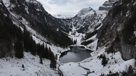 Aerial-view-of-Tahlalpsee-in-Filzbach,-Glarus-Nord,-Switzerland,-cradled-by-snow-covered-mountains-and-alpine-forests