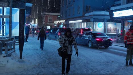 Night-time-camera-shot-showing-traffic-marshal-directing-car-traffic-into-the-resort-with-snow-falling,-pedestrians-walking-and-bright-billboards