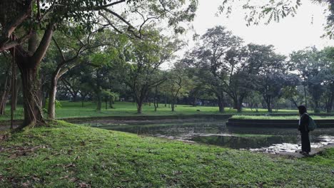 Wide-panoramic-view-of-a-young-female-student-wearing-backpack-looking-across-idyllic-nature-park-with-large-trees-and-lake-in-campus