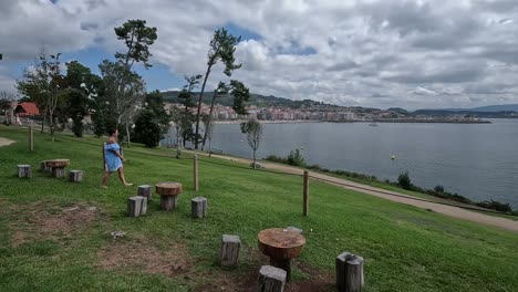 Woman-goes-out-to-enjoy-the-natural-grass-terrace-with-rustic-wooden-seats-and-tables-facing-the-sea-and-the-city-is-seen-in-the-background,-cloudy-day,-descriptive-shot