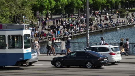 Toma-Lenta-De-Un-Dron-En-Movimiento-Hacia-La-Izquierda-Con-Un-Tranvía-De-Zurich-Moviéndose-Hacia-La-Izquierda-Fuera-Del-Marco,-Revelando-Multitudes-Sentadas-Frente-Al-Lago-De-La-Ciudad.