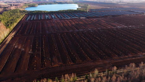 Aerial-establishing-shot-of-peat-extraction-field-rows-on-edge-of-forest-by-lake